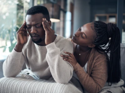 Mulher consolando companheiro no sofá, abraçada nele. Ele está tocando as têmporas, com feições de dor.